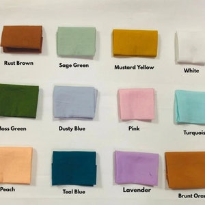 May include: An assortment of folded fabric swatches in various colors, including rust brown, sage green, mustard yellow, and white. Additional colors are moss green, dusty blue, pink, turquoise, peach, teal blue, lavender, and burnt orange. Each swatch has a color name printed below it.