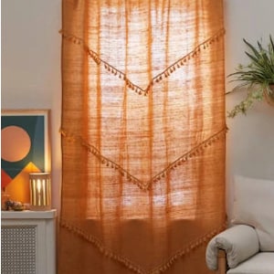 May include: A burnt orange curtain with a textured weave hangs in front of a window. The curtain features a scalloped top and decorative chevron patterns with small tassels. The curtain is suspended from a gold-colored rod.