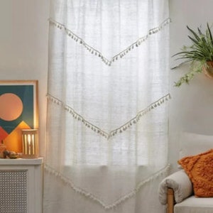 May include: White linen curtain with a gold rod. The curtain features three chevron-shaped rows of tassels. The top of the curtain has a scalloped edge. A cozy living room setting with a chair and artwork is visible.