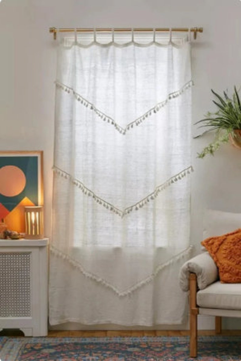 May include: White linen curtain with a gold rod. The curtain features three chevron-shaped rows of tassels. The top of the curtain has a scalloped edge. A cozy living room setting with a chair and artwork is visible.
