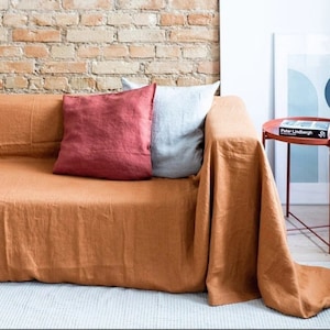 以下が含まれることがあります： レンガの壁の前に、2つのクッション（赤とグレー）を置いた茶色のリネン製のソファカバー。ソファカバーはソファの背もたれにかけられ、床まで垂れ下がっています。背景には、「ピーター・リンドバーグ」というタイトルの本が置かれた小さな赤い金属製のサイドテーブルがあります。