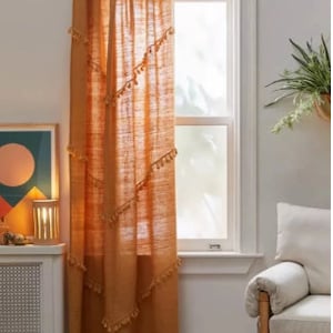 May include: A burnt orange linen curtain hangs in front of a window. The curtain is adorned with small tassels along diagonal lines. A gold-colored curtain rod is at the top. The window is framed by white trim. A framed piece of art and a lamp are visible.