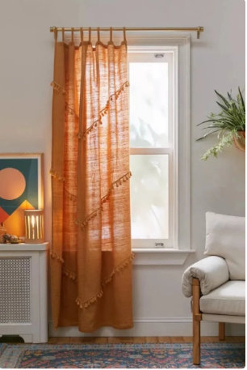 May include: A burnt orange linen curtain hangs in front of a window. The curtain is adorned with small tassels along diagonal lines. A gold-colored curtain rod is at the top. The window is framed by white trim. A framed piece of art and a lamp are visible.