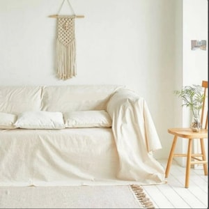 May include: A cream-colored sofa covered with a matching fabric throw, featuring three square cushions. Above the sofa hangs a cream macrame wall hanging with a wooden dowel. To the right, a light wood chair holds a small glass vase with greenery.