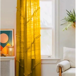 May include: Mustard yellow curtain panel with decorative tassel trim, hanging from a gold-colored rod. The curtain is sheer, allowing light to filter through. The window is framed by white trim, and the room has a bohemian aesthetic.