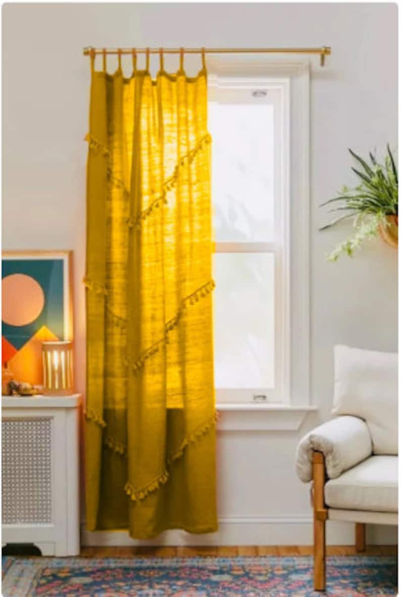 May include: Mustard yellow curtain panel with decorative tassel trim, hanging from a gold-colored rod. The curtain is sheer, allowing light to filter through. The window is framed by white trim, and the room has a bohemian aesthetic.