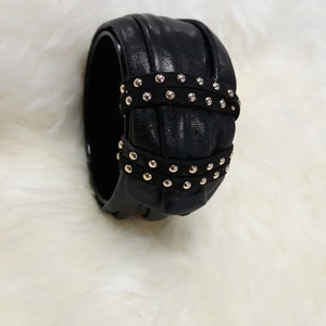 Puede incluir: Pulsera de cuero negro con tachuelas doradas. La pulsera es ancha y tiene una superficie texturizada.