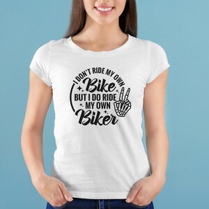 May include: White t-shirt with a black graphic that reads "I don't ride my own bike but I do ride my own biker" with a skeleton hand making a peace sign.