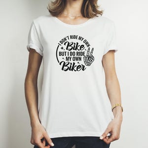 May include: White t-shirt with a black graphic that says "I don't ride my own bike, but I do ride my own biker". The graphic also includes a skeleton hand giving the peace sign.