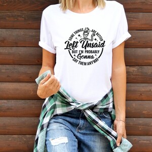 May include: White t-shirt with a black and white graphic design. The graphic features a skeleton holding a drink and the text "Some things are better Left Unsaid. But I'm probably gonna say them anyway."
