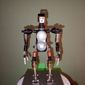 May include: A handcrafted robot sculpture made from recycled metal and electronic parts. The robot has a black body with silver and copper details, articulated arms and legs, and a shaver head.