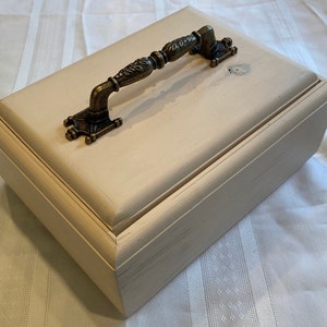 May include: A white wooden box with a decorative brass handle. The box has a slightly distressed finish and a raised edge around the lid.