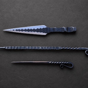 May include: Three hand-forged metal tools arranged horizontally. The top item is a wide, pointed blade with a textured edge. The middle item is a long, thin rod with a notched surface and a curled end. The bottom item is a slender, pointed pick.