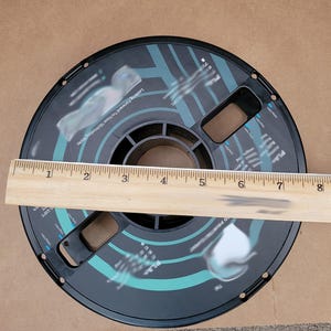 May include: A black spool of filament for a 3D printer. The spool is 8 inches in diameter and has a blue and black design. A wooden ruler is placed on top of the spool for scale.