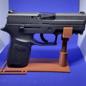 May include: A black handgun rests on a brown stand with a threaded support. The gun has a textured grip and a metallic slide. The stand is rectangular with a cutout design and a vertical support.