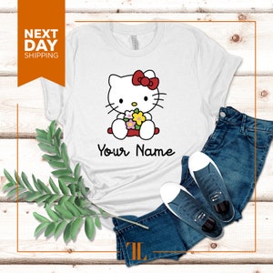 May include: White t-shirt with a cartoon image of Hello Kitty holding flowers. The text "Your Name" is printed below the image.