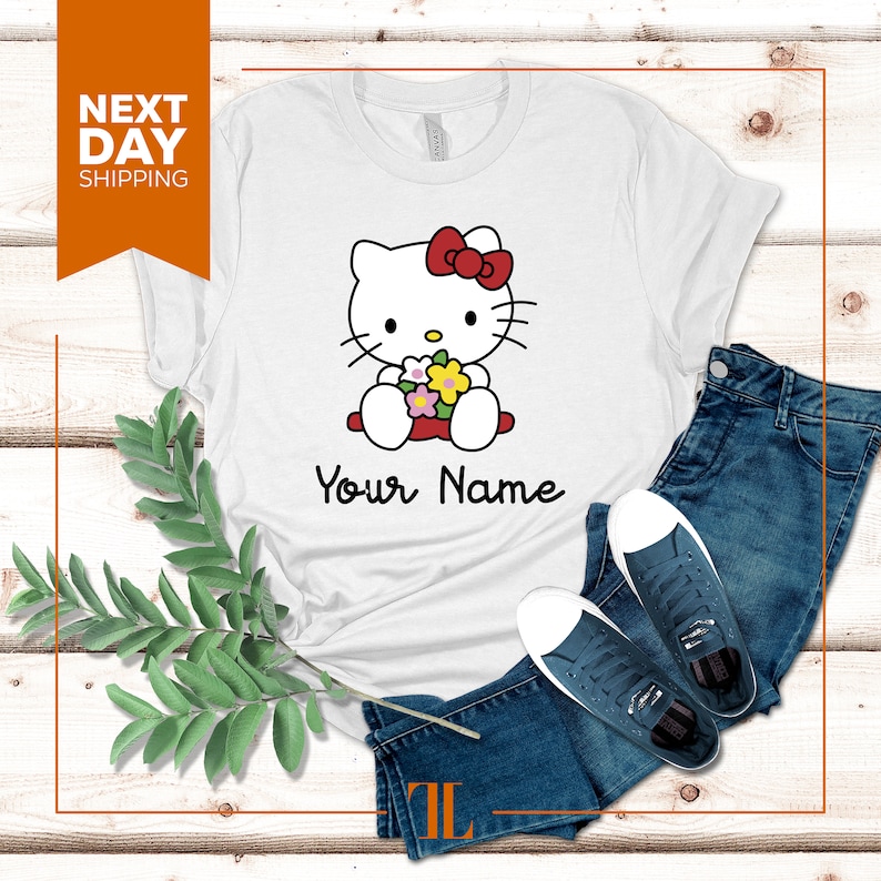 May include: White t-shirt with a cartoon image of Hello Kitty holding flowers. The text "Your Name" is printed below the image.