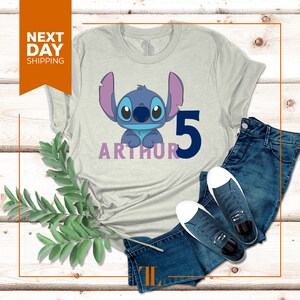May include: A light gray t-shirt with a blue Stitch character from Lilo & Stitch. The shirt has the name "Arthur" and the number "5" printed on it.