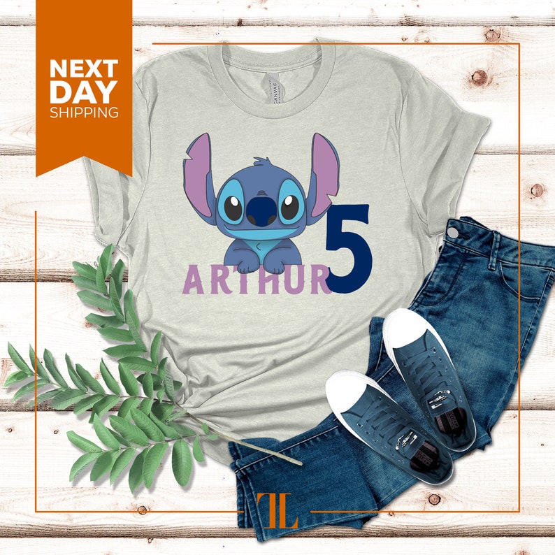May include: A light gray t-shirt with a blue Stitch character from Lilo & Stitch. The shirt has the name "Arthur" and the number "5" printed on it.