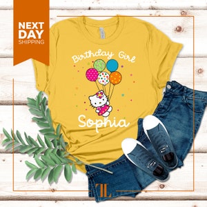 Puede incluir: Camiseta amarilla con un gr&aacute;fico de un gato de dibujos animados sosteniendo globos que dicen "Birthday Girl" y el nombre "Sophia".