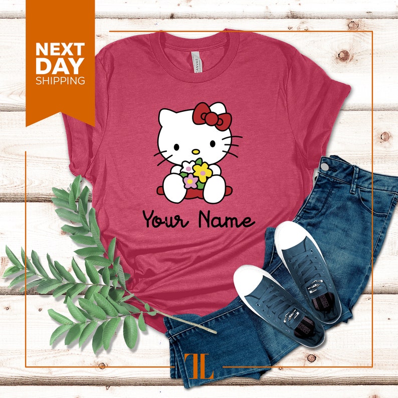 May include: A red t-shirt with a white Hello Kitty graphic holding flowers. The text "Your Name" is printed below the graphic.