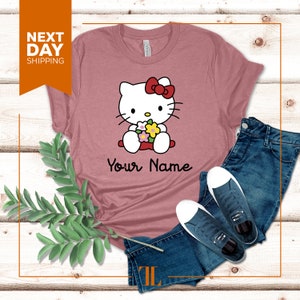 May include: A pink t-shirt with a cartoon image of Hello Kitty holding flowers. The text "Your Name" is printed below the image.