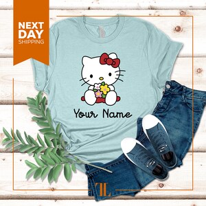 May include: Light blue t-shirt with a cartoon image of Hello Kitty holding flowers. The text "Your Name" is printed below the image.