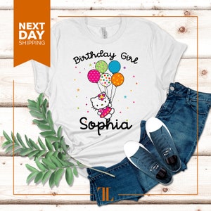 Puede incluir: Camiseta blanca con un dise&ntilde;o de ni&ntilde;a de cumplea&ntilde;os con un gato de dibujos animados sosteniendo globos. El texto "Birthday Girl" y "Sophia" est&aacute; impreso en la camiseta.