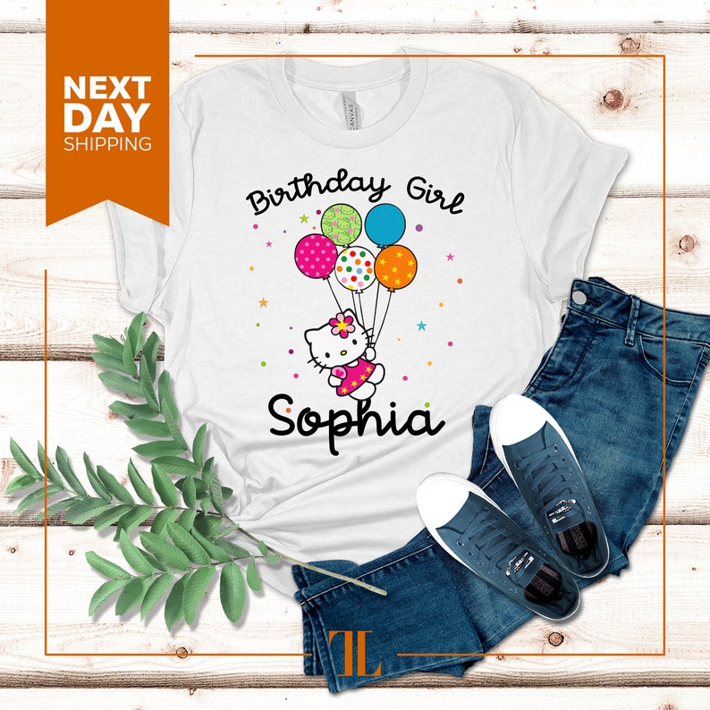 Puede incluir: Camiseta blanca con un dise&ntilde;o de ni&ntilde;a de cumplea&ntilde;os con un gato de dibujos animados sosteniendo globos. El texto "Birthday Girl" y "Sophia" est&aacute; impreso en la camiseta.