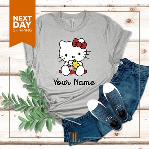 May include: Gray t-shirt with a cartoon image of Hello Kitty holding flowers. The text "Your Name" is printed below the image.