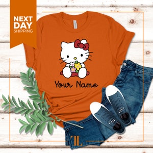 May include: Orange t-shirt with a cartoon image of Hello Kitty holding flowers. The text "Your Name" is printed below the image.