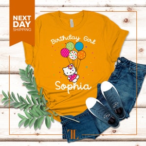 Puede incluir: Camiseta amarilla con un dise&ntilde;o de ni&ntilde;a de cumplea&ntilde;os con un personaje de dibujos animados sosteniendo globos. El texto "Birthday Girl" y "Sophia" est&aacute;n impresos en la camiseta.