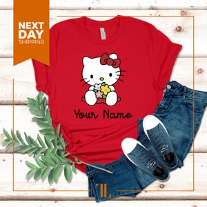 May include: Red t-shirt with a cartoon image of Hello Kitty holding flowers. The text "Your Name" is printed below the image.