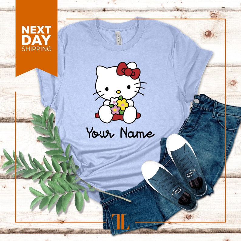 May include: Light blue t-shirt with a cartoon image of Hello Kitty holding flowers. The text "Your Name" is printed below the image.