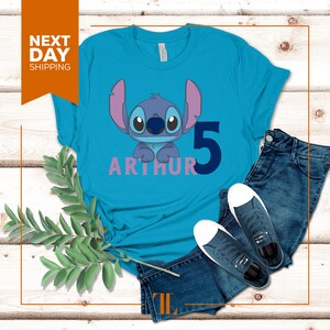 May include: A turquoise blue t-shirt with a blue Stitch character from Lilo & Stitch. The shirt has the name "Arthur" and the number "5" printed on it in pink and blue.