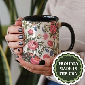May include: A ceramic mug with a black handle and interior, featuring a cream-colored exterior decorated with a colorful floral pattern of pink roses, tulips, and daisies. A green scalloped badge on the mug reads PROUDLY MADE IN THE USA.