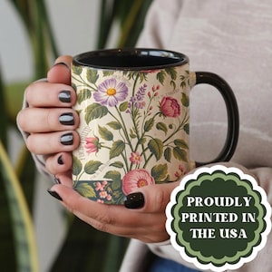 May include: A ceramic mug with a black interior and handle, featuring a cream-colored exterior decorated with a botanical illustration of pink, purple, and white flowers and green leaves. The text FUCK TRUMP is integrated into the floral design.