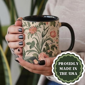 May include: A ceramic mug with a black handle and interior, featuring a cream-colored exterior decorated with a botanical pattern of flowers and green leaves. The text "FUCK TRUMP" is printed in small, repeating script throughout the floral design.