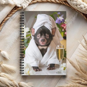 May include: A black Tasmanian devil wearing a white bathrobe and a white towel on its head, sitting on a white surface with a glass of champagne next to it.