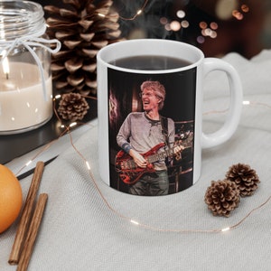May include: White ceramic mug with a black and white image of a man playing a red electric guitar. The man is wearing a grey sweater and has a beard.