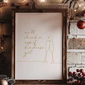 May include: A framed print with a gold inscription that reads "we'll drink a cup of kindness yet" with a simple line drawing of two champagne glasses clinking. The print is set against a brick wall with Christmas lights and ornaments.