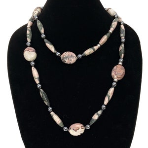May include: A long necklace featuring a mix of oval and cylindrical beads in shades of pink, gray, and black. The necklace is accented with small, silver-colored beads and is displayed on a black mannequin.