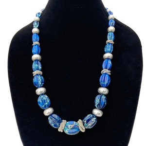 May include: A blue beaded necklace with silver accents. The necklace features large, oval beads in shades of blue, interspersed with patterned silver beads. The necklace is displayed on a black mannequin.