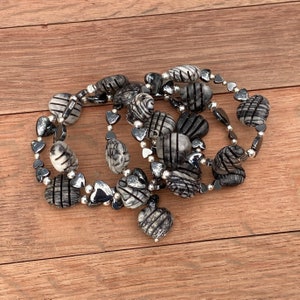 Puede incluir: Un collar con una mezcla de cuentas a rayas y estampadas en blanco y negro, intercaladas con cuentas plateadas en forma de corazón y redondas. El collar se exhibe sobre una superficie de madera.