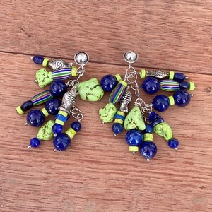 Op de afbeelding: Een paar oorhangers met blauwe, groene en zilveren elementen. De oorbellen hebben een zilveren knop, met bungelende kettingen met blauwe kralen, groene stenen en zilveren visbedels. De oorbellen zijn geplaatst tegen een houten achtergrond.