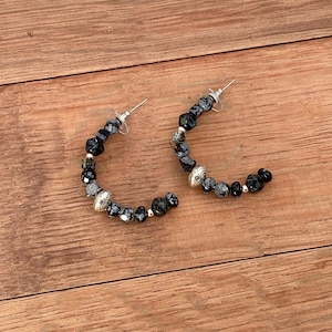 May include: A pair of hoop earrings with a post back. The earrings are made of black and gray faceted beads and silver-colored metal beads. The earrings are displayed on a wooden surface.