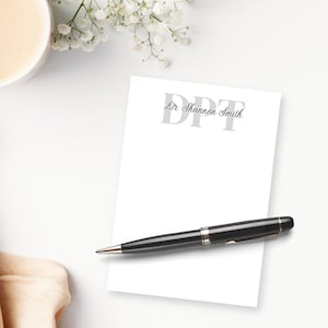 May include: A white notepad with the text "DPT Dr. Shannon Smith" in gray lettering. A black pen with silver accents rests on the notepad. The scene includes a cup of coffee and white flowers.