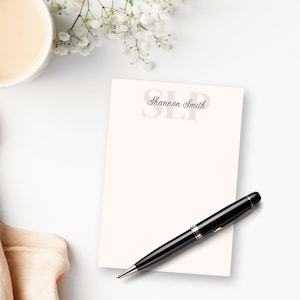 May include: A white notepad with a pink background and the text "Shannon Smith SLP" in black script. A black pen is lying on the notepad.