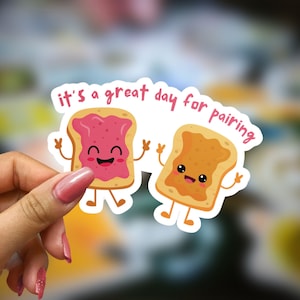 May include: Two cartoon slices of bread, one with strawberry jam and one with peanut butter, with the text "it's a great day for pairing".