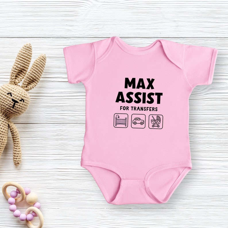 Może przedstawiać: R&oacute;żowy śpioszek dla niemowlęcia z czarnym napisem "Max Assist for Transfers" i trzema ikonami: ł&oacute;żeczkiem, samochodem i w&oacute;zkiem.